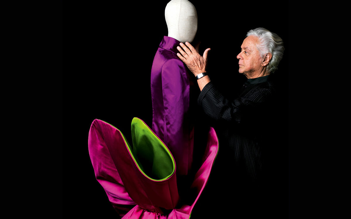 Roberto Capucci in piedi accanto a un grande abito scultoreo in raso disposto a cerchio.